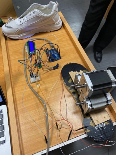 Photograph of the physical smart shoe prototype setup. The image shows a white sneaker connected to an array of wires, an Arduino board, an LCD screen, and a mechanical actuator, all mounted on a wooden board. This setup demonstrates the integration of sensors, electronics, and mechanical components used for real-time gait and pressure measurement.