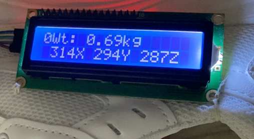 Close-up photo of the smart shoe's LCD screen display. The screen shows weight measurement in kilograms (e.g., '0.69kg') and three numerical values labeled X, Y, and Z, representing foot orientation or acceleration data. This display provides real-time feedback to the user about their weight and foot movement.